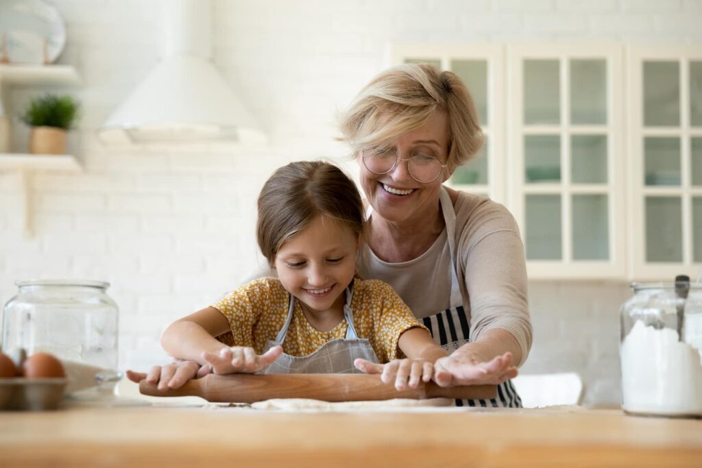 A grandmother and grandchild roll out dough together in a kitchen.
