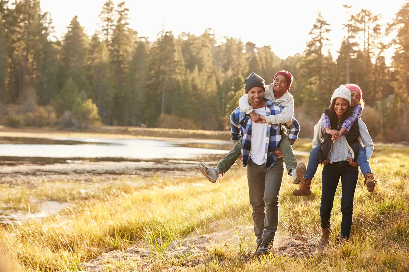 Parents walk with their children on their backs through the outdoors near a lake.